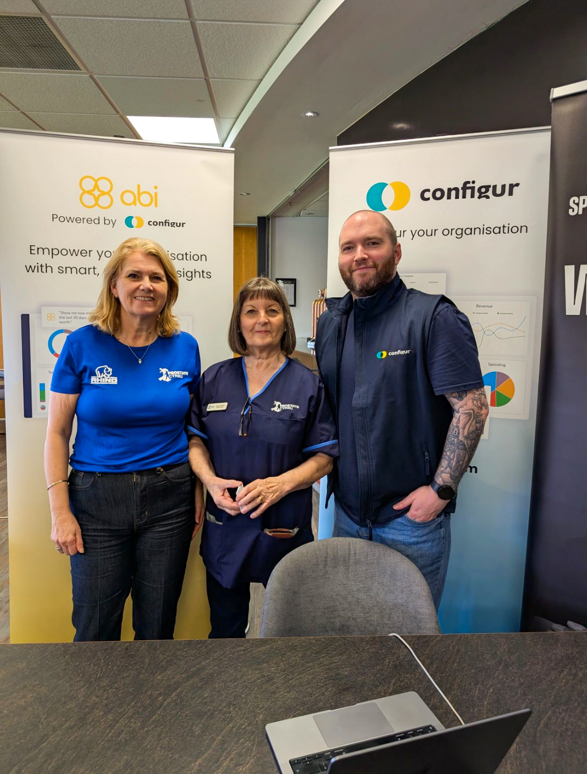A group of three people posing for a photo in front of promotional banners for abi powered by configur and configur, with a laptop and a table visible in the foreground.